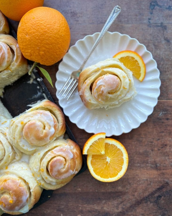 An orange sweet roll on a plate with oranges around it.