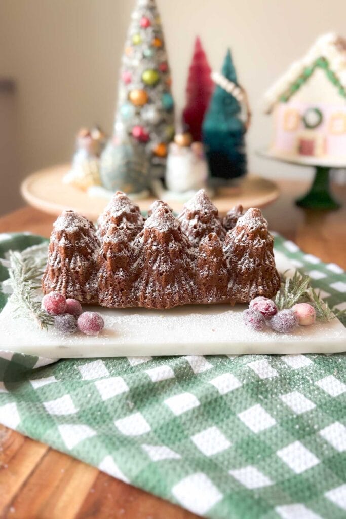 A loaf of spiced gingerbread on a green and white checked towel with Christmas decorations in the background.